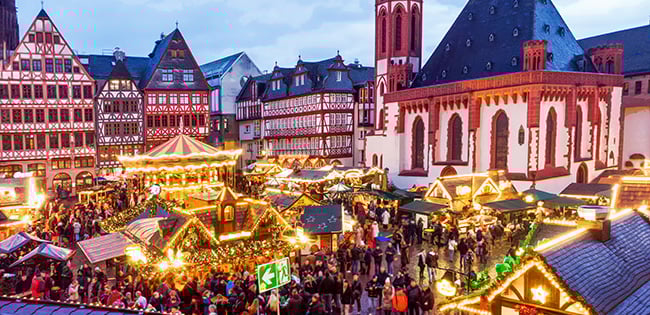 A lively Christmas market in Germany, set against a backdrop of historic half-timbered buildings and a grand church. The market is illuminated with festive lights, and the bustling crowd enjoys the decorated stalls offering holiday treats, crafts, and gifts under the evening sky.