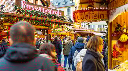 A bustling scene from a German Christmas market, where people gather around festive wooden stalls adorned with holiday decorations. Signs for 'Feuerzangenbowle' and 'Waffeln' highlight the traditional seasonal treats available, with warm lights and greenery enhancing the cozy atmosphere.