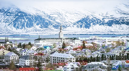 A picturesque view of Reykjavik, Iceland, featuring colorful rooftops and charming buildings against a backdrop of snow-covered mountains. The serene landscape includes a glimpse of water in the distance, adding to the city's tranquil atmosphere. The bright, wintry scene captures the unique blend of urban and natural beauty characteristic of Iceland.