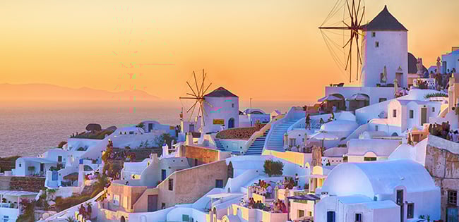 A stunning sunset view of Oia, Santorini, Greece, featuring iconic whitewashed buildings with blue domes cascading down a hillside toward the sea. Traditional windmills stand prominently against the warm orange and pink hues of the sky. The Aegean Sea glows softly in the fading light, creating a serene and picturesque scene.