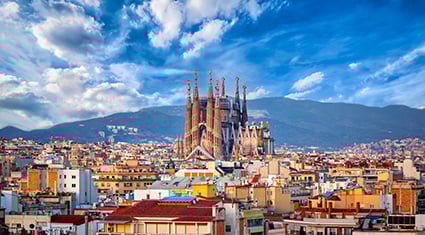 A breathtaking view of Barcelona, Spain, featuring the iconic Sagrada Família basilica rising prominently above the cityscape. The colorful rooftops of surrounding buildings create a vibrant foreground, while rolling hills and a dramatic sky filled with clouds add depth and contrast to the scene.