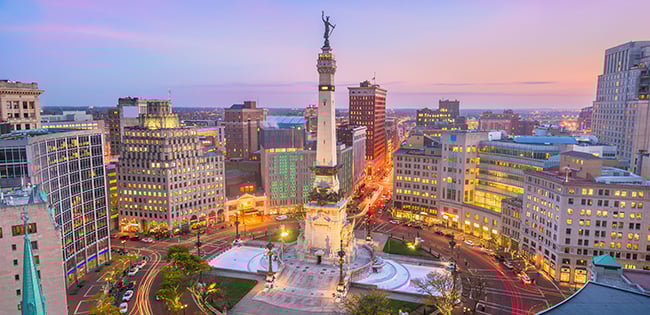 A vibrant evening view of Monument Circle in Indianapolis, Indiana. The Soldiers and Sailors Monument stands prominently in the center, surrounded by illuminated buildings and streets radiating out in a circular pattern. The sky is painted with hues of purple, pink, and orange as the city transitions into night, creating a lively and picturesque urban scene.