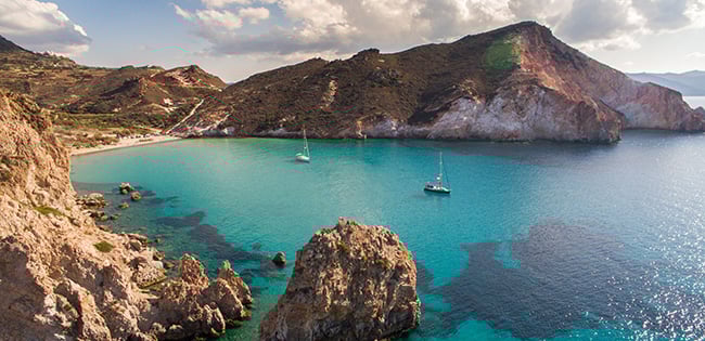 A stunning view of a secluded bay in the Greek Isles, featuring turquoise waters surrounded by rugged cliffs and rocky formations. Sailboats are anchored in the calm water, and the shoreline has a pristine, sandy beach. The scene is illuminated under a partly cloudy sky, highlighting the serene and natural beauty of the Mediterranean landscape.