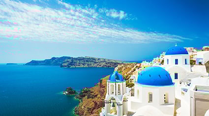 A picturesque view of Santorini, Greece, featuring iconic whitewashed buildings with blue domes perched on a cliff overlooking the deep blue Aegean Sea. The coastline extends into the distance, framed by a vibrant blue sky with wispy clouds. The stunning contrast of colors highlights the charm and beauty of this famous Mediterranean destination.