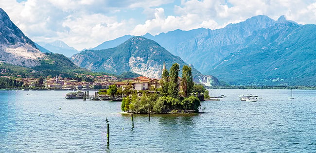 A picturesque view of Lake Maggiore in Italy, featuring a small island with lush greenery and historic buildings. The lake is surrounded by majestic mountains, creating a serene and scenic backdrop. Boats are scattered across the calm waters, and the distant shoreline is dotted with charming towns under a partly cloudy blue sky.