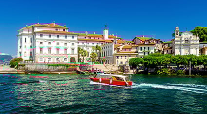 A vibrant waterfront scene of Isola Bella on Lake Maggiore in Italy, showcasing elegant white buildings with red-tiled roofs and lush greenery. A small red and white boat cruises across the sparkling blue water, adding movement to the serene setting. The bright blue sky enhances the charm and beauty of this picturesque Italian destination.