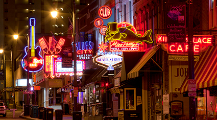 A vibrant nighttime scene of Beale Street in Memphis, Tennessee, showcasing colorful neon signs for blues clubs, restaurants, and shops, including a guitar-shaped sign and a bright fish wearing a top hat. The street captures the lively energy of Memphis' famous music and entertainment district.