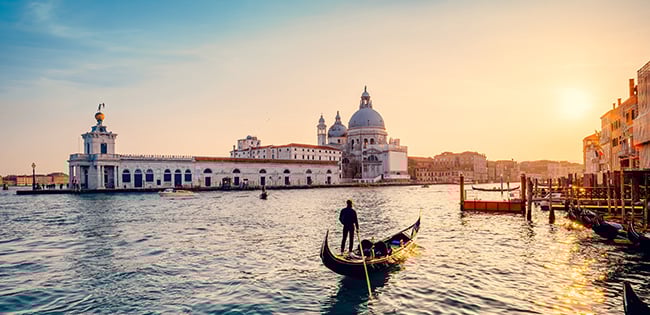 A serene view of Venice, Italy, at sunset, featuring a gondolier navigating a gondola across the calm waters of the Grand Canal. The iconic Santa Maria della Salute basilica stands prominently in the background, bathed in the warm glow of the setting sun. Historic buildings line the canal, adding to the romantic and timeless atmosphere of the scene.