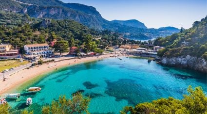 A scenic beach cove in Corfu, Greece, with crystal-clear turquoise water, small boats anchored near the shore, sandy beach, and lush green hills rising in the background.