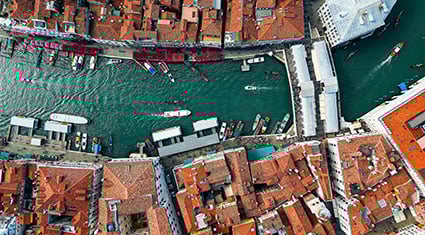 An aerial view of Venice, Italy, showcasing the Grand Canal winding through the city. The scene is framed by historic buildings with red-tiled roofs, and the canal is dotted with boats, gondolas, and water taxis. Bridges connect the sides of the canal, adding to the charm of this iconic Venetian landscape. The vibrant colors and unique layout highlight the city's timeless beauty.