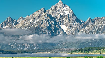 A stunning view of the Grand Teton mountain range, featuring a jagged, snow-capped peak rising dramatically against a bright blue sky. The foreground showcases a serene lake surrounded by lush green meadows and forests. Low-lying clouds add a soft, atmospheric touch to the breathtaking landscape.