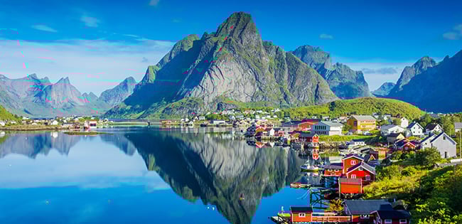 A breathtaking view of a small fishing village in the Lofoten Islands, Norway, featuring traditional red and white houses along a calm, reflective fjord. Towering green mountains rise dramatically in the background under a bright blue sky. The vibrant colors and serene atmosphere highlight the natural beauty and charm of this Nordic destination.