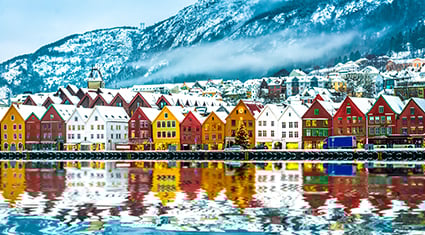 A stunning winter view of the historic Bryggen district in Bergen, Norway, featuring colorful wooden houses in red, yellow, and white along the waterfront. The buildings are reflected in the calm water, and the surrounding mountains are dusted with snow and partially shrouded in mist, creating a picturesque and serene atmosphere.