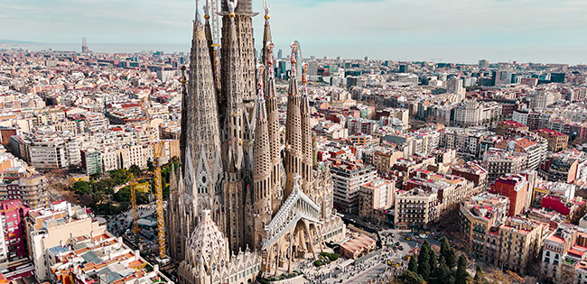 An aerial view of the iconic Sagrada Família basilica in Barcelona, Spain, showcasing its intricate spires and Gothic-inspired architecture surrounded by a bustling cityscape of buildings and streets. The detailed craftsmanship of the basilica contrasts beautifully with the modern urban environment.