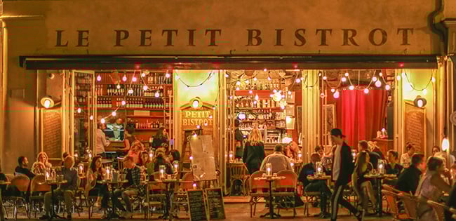 A cozy evening scene of "Le Petit Bistrot" in Provence, France. The warmly lit café features outdoor seating with small tables and chairs, where patrons enjoy food and drinks. The open storefront showcases shelves of wine bottles and a welcoming ambiance. String lights and soft red curtains add to the charming atmosphere, with people socializing and savoring the relaxed vibe.