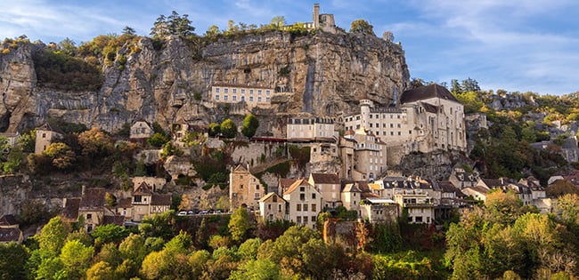 A picturesque view of Rocamadour, France, a medieval village built into the side of a steep cliff. The stone buildings and a historic church are nestled among the rugged rock formations, surrounded by lush greenery and autumn-colored trees. A bright blue sky with scattered clouds adds to the charm and dramatic beauty of this unique hillside village.