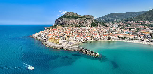 A breathtaking aerial view of Cefalù, a coastal town in Sicily, Italy. The picturesque town features tightly packed, terracotta-roofed buildings nestled along the shoreline, with a prominent medieval cathedral standing out near the center. A rugged cliff rises dramatically behind the town, while the turquoise waters of the Tyrrhenian Sea surround the peninsula. A small boat leaves gentle ripples in the water, adding a sense of movement to the serene landscape.