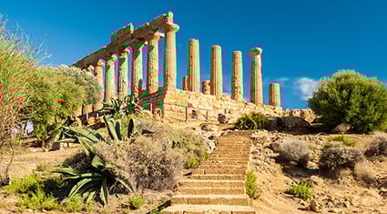 An iconic ancient Greek temple located in the Valley of the Temples in Agrigento, Sicily, Italy. The temple features well-preserved Doric columns, basking in the warm glow of sunlight. The surrounding area is dotted with lush greenery, including cacti and Mediterranean shrubs, set against a backdrop of a clear blue sky. A stone pathway and steps lead up to the historic structure, enhancing its grandeur and accessibility.