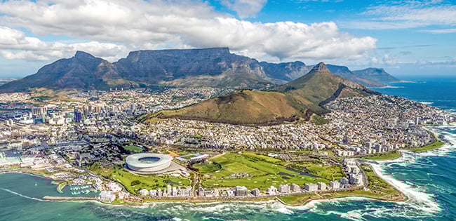 A breathtaking aerial view of Cape Town, South Africa, showcasing the city's vibrant urban landscape surrounded by the ocean. Iconic landmarks such as Table Mountain and Lion's Head rise majestically in the background under a bright blue sky with scattered clouds. The coastline features rugged rocks and white waves, with the Cape Town Stadium visible near the shoreline.