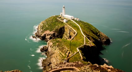 A scenic view of the South Stack Lighthouse perched atop a rugged green peninsula on the coast of Anglesey, Wales. A winding footpath leads across steep cliffs to the white lighthouse, surrounded by calm, turquoise waters.