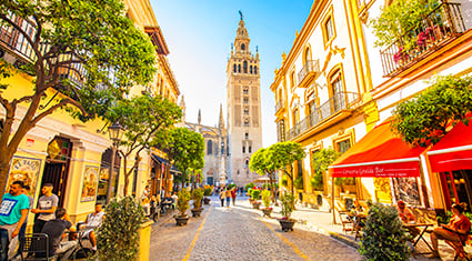 A lively street scene in Seville, Spain, featuring the Giralda bell tower in the background and people dining at outdoor cafes beneath colorful buildings.
