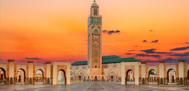 The Hassan II Mosque in Casablanca, Morocco, stands prominently at sunset, with its towering minaret and intricate architecture framed by a courtyard of arches under a vivid orange and purple sky.