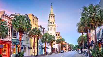 A charming street view in Charleston, South Carolina, lined with palm trees and pastel-colored historic buildings with a church steeple in the distance.