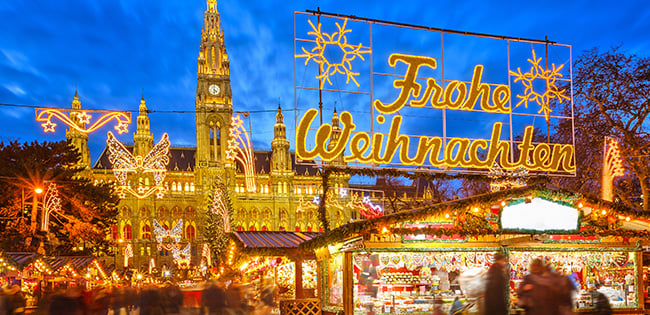 Vienna’s Rathausplatz glows during the Christmas market, with festive lights, decorated stalls, and a large “Frohe Weihnachten” sign illuminating the square.