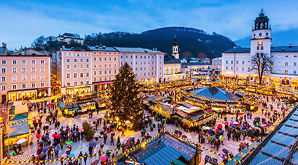 A bustling Christmas market fills the historic town square of Salzburg, Austria, with twinkling lights, holiday decorations, and a large Christmas tree.
