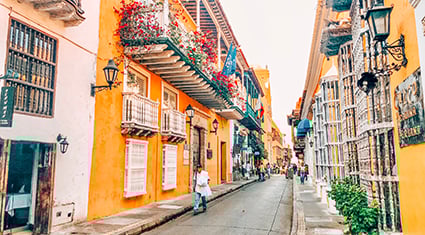 A colorful colonial street in Cartagena, Colombia, with orange-washed buildings, flower-filled balconies, and pedestrians strolling along the cobblestones.