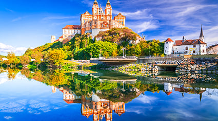 Melk Abbey in Austria stands majestically on a hill above the Danube River, with its baroque architecture reflecting in the calm water below.