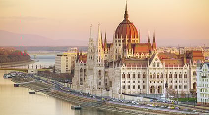 The Hungarian Parliament Building along the Danube River in Budapest glows under the soft light of sunset, with its spires and dome dominating the riverbank.