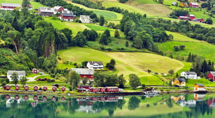 Picturesque Norwegian countryside with colorful houses and farms reflected in a calm fjord, framed by rolling green hills and dense forest.