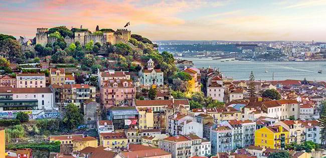 A panoramic view of Lisbon, Portugal, featuring São Jorge Castle on a hill surrounded by colorful buildings and the Tagus River in the background.