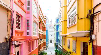 A narrow, charming alleyway in A Coruña, Spain, lined with bright pink, yellow, and blue buildings, leading to a cathedral in the distance.