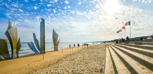 View of Omaha Beach in Normandy, France, featuring the Les Braves D-Day memorial sculpture, national flags and visitors walking along the sandy shore under a bright sky.