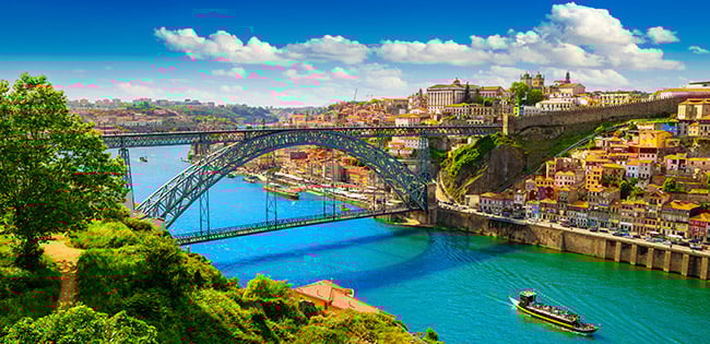 The Dom Luís I Bridge spans the Douro River in Porto, Portugal, connecting the colorful cityscapes on either side under a bright blue sky.