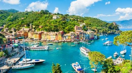 Colorful waterfront buildings line the harbor of Portofino, Italy, surrounded by lush green hills, with sailboats and yachts floating in the turquoise Ligurian Sea.