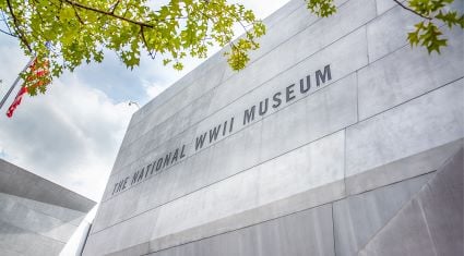 Close-up of The National WWII Museum’s modern concrete facade with bold lettering, framed by leafy green branches against a partly cloudy sky.