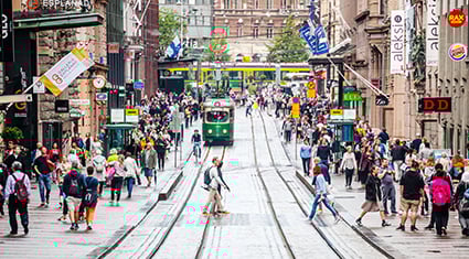 A bustling street scene in Helsinki, Finland, with trams, shops, and pedestrians moving along a vibrant city thoroughfare.