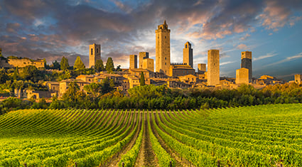 The medieval towers of San Gimignano, Italy, rise above lush vineyards in the golden light of sunset, creating a dramatic skyline.