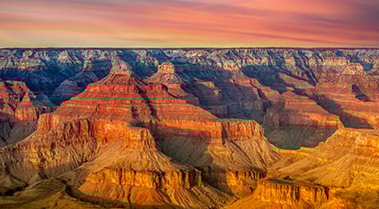 A vivid sunset casts golden light across the layered rock formations of the Grand Canyon, highlighting its vast, colorful expanse.