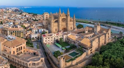 Aerial view of Palma de Mallorca, Spain, featuring the Gothic-style La Seu Cathedral near the coastline, surrounded by historic buildings, tree-lined streets, and the sparkling blue Mediterranean Sea.