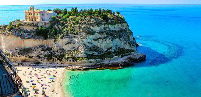 A scenic beach in Tropea, Italy, with turquoise waters and sunbathers at the base of a rocky cliff topped by the Sanctuary of Santa Maria dell’Isola.