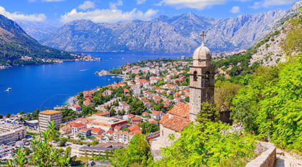The Bay of Kotor in Montenegro, with red-roofed buildings nestled between the mountains and sea, and a stone church tower rising in the foreground.