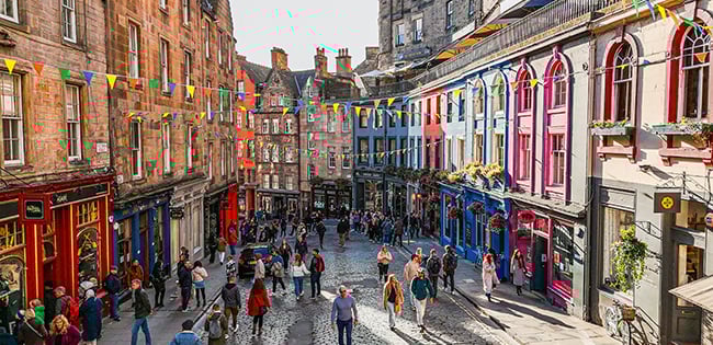 A lively street scene in Edinburgh, Scotland, featuring colorful storefronts on Victoria Street with people walking beneath festive pennant flags.