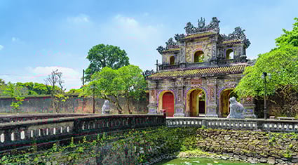The ornate Ngo Mon Gate at the Imperial City in Hue, Vietnam, with red and gold detailing, flanked by stone lions and shaded by trees.