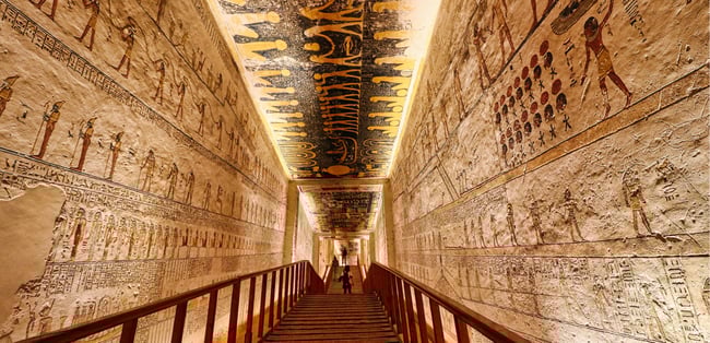 An ornately decorated passageway inside an ancient Egyptian tomb, featuring vibrant hieroglyphics and figures etched and painted on the walls and ceiling. Warm lighting illuminates the detailed artwork as visitors descend the wooden steps into the historic burial chamber.