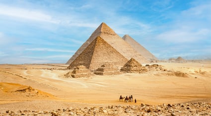 A wide-angle view of the iconic Pyramids of Giza rising from the golden sands of the Egyptian desert. Several travelers on camels can be seen in the foreground, emphasizing the vast scale and ancient grandeur of the surrounding ruins under a bright blue sky.