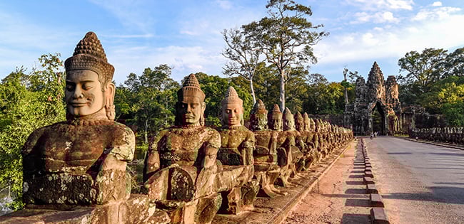 A line of stone statues depicting deities and demons lines the entrance bridge to Angkor Thom in Cambodia, with the South Gate visible in the background.